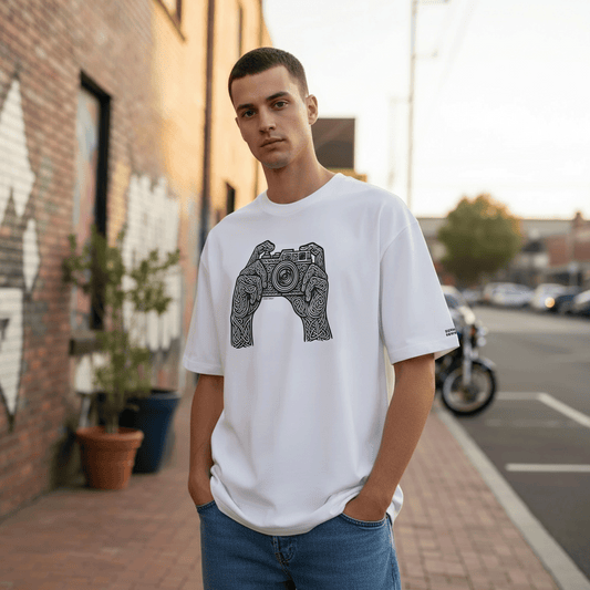 Model standing on a quiet street wearing a white box fit tee with carved black and white camera artwork while the evening light softens the background.