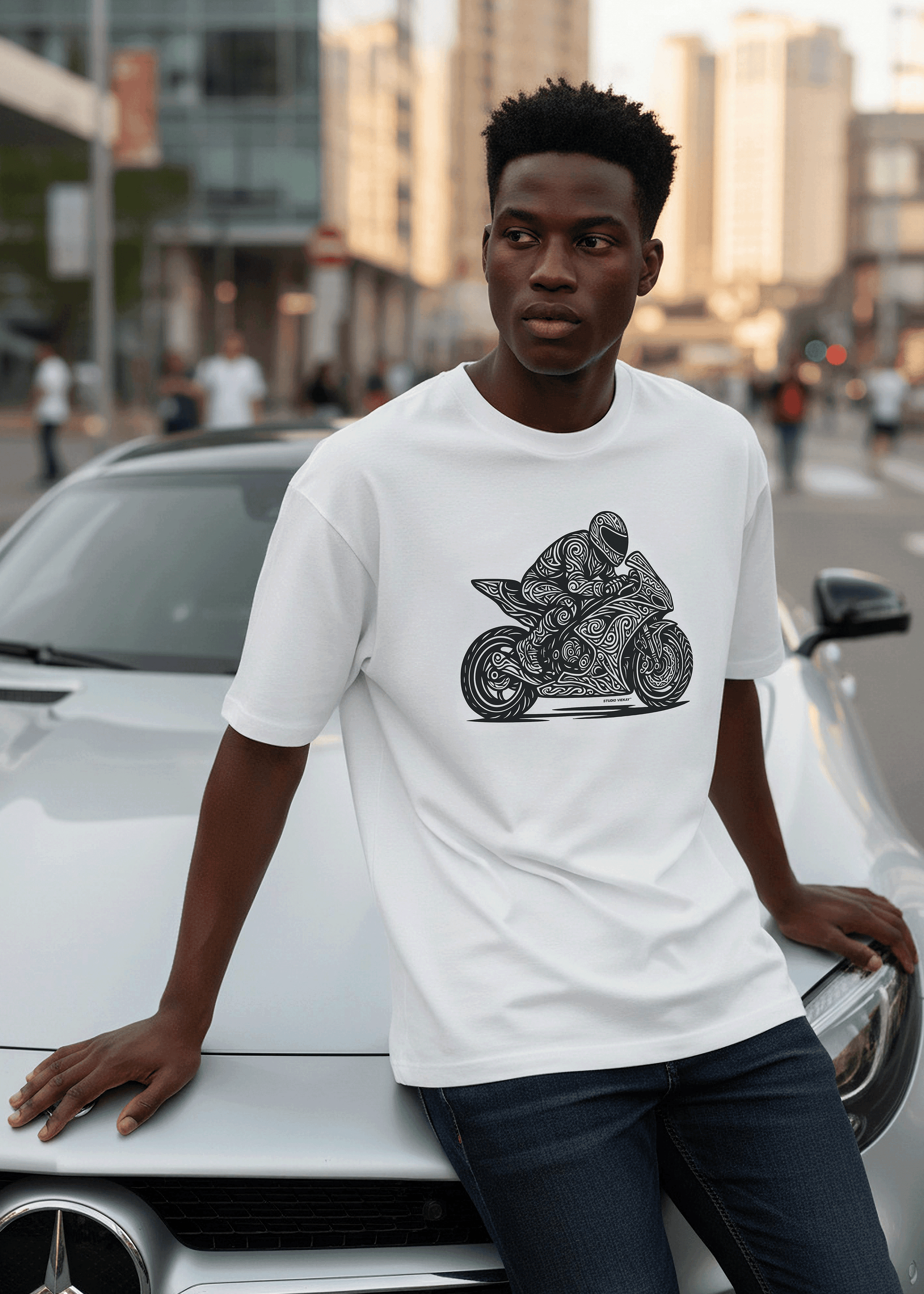 Model wearing Studio Viekay Motorcycle Engraved Art Box Tee while leaning against a white car in an urban street setting