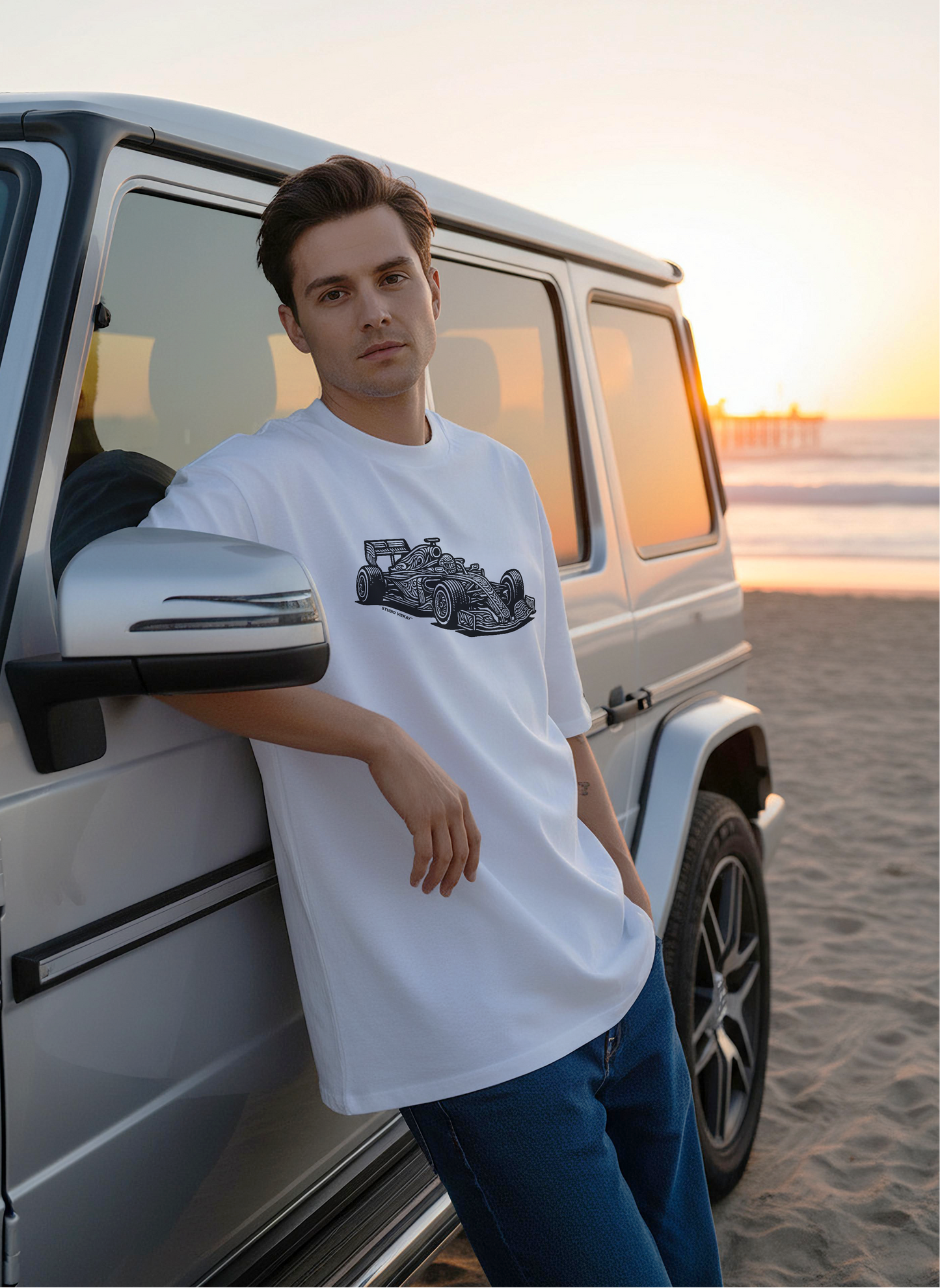Model leaning against a silver SUV on the beach at sunset wearing a white box fit tee with carved black and white Formula One artwork.