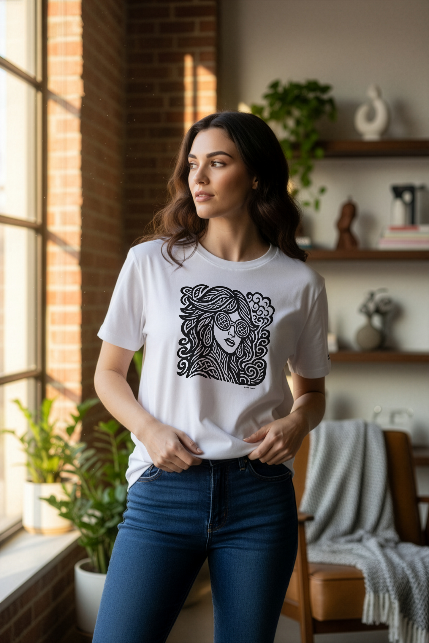 Model wearing a white Studio Viekay t-shirt with a black graphic design in a room with plants and shelves.