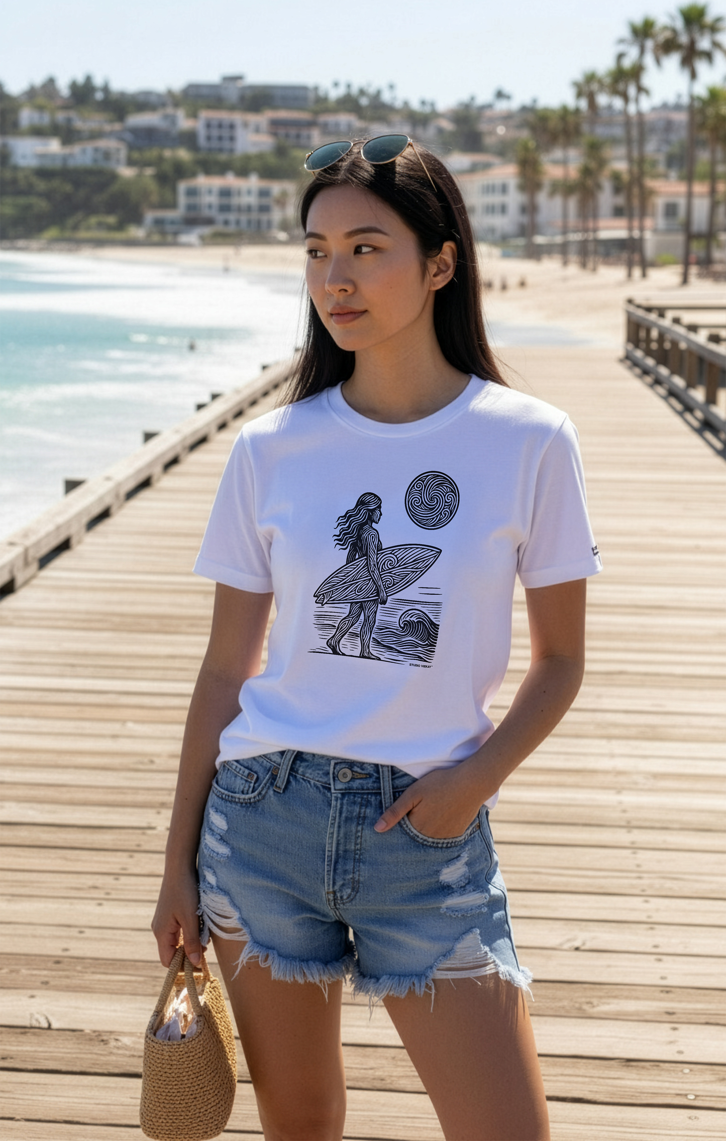 Model wearing a white Studio VieKay t-shirt with a graphic design on a wooden pier by the beach.
