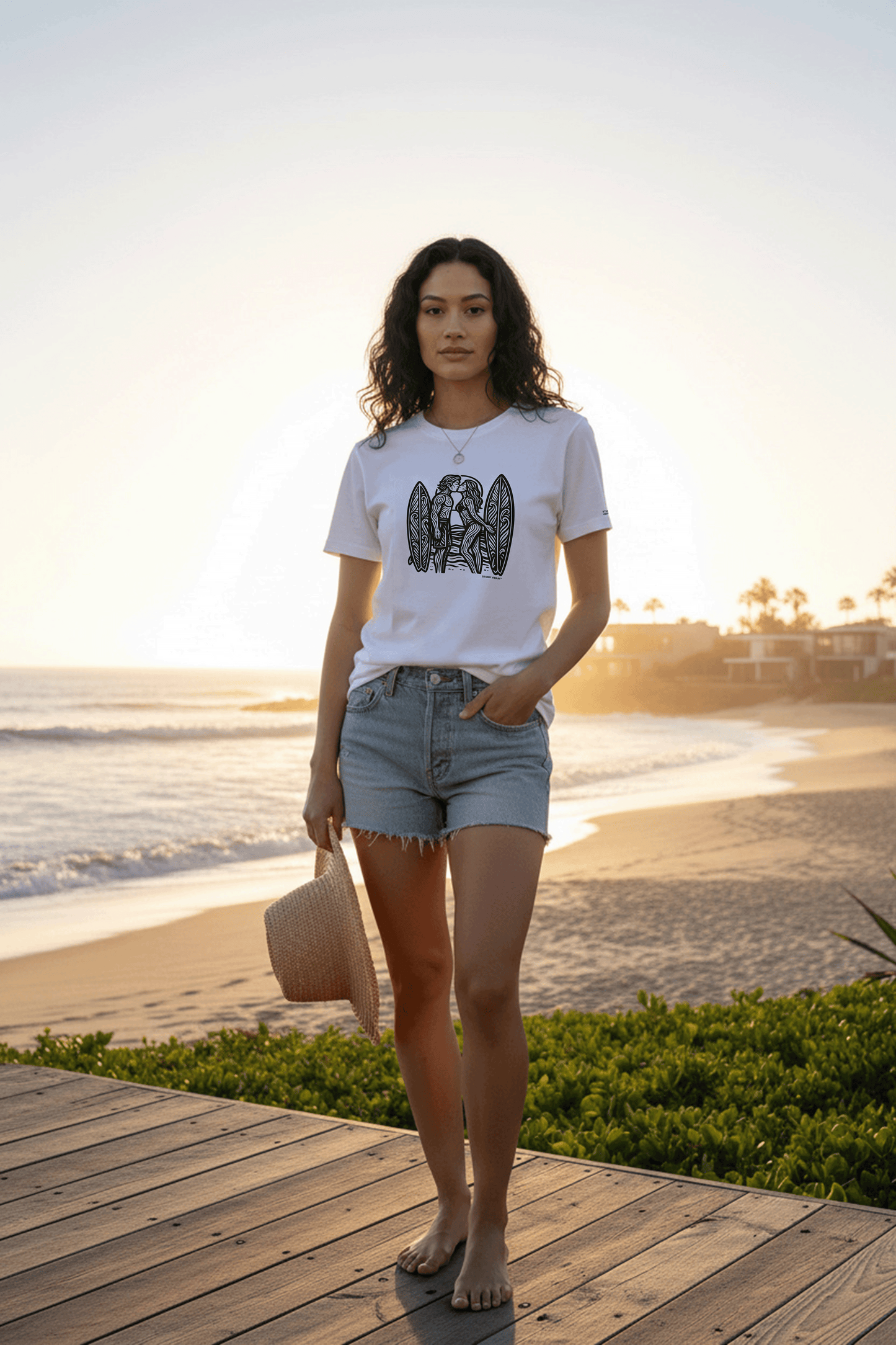 Model standing on a wooden deck by the beach at sunset, wearing a graphic tee by Studio Viekay and denim shorts.