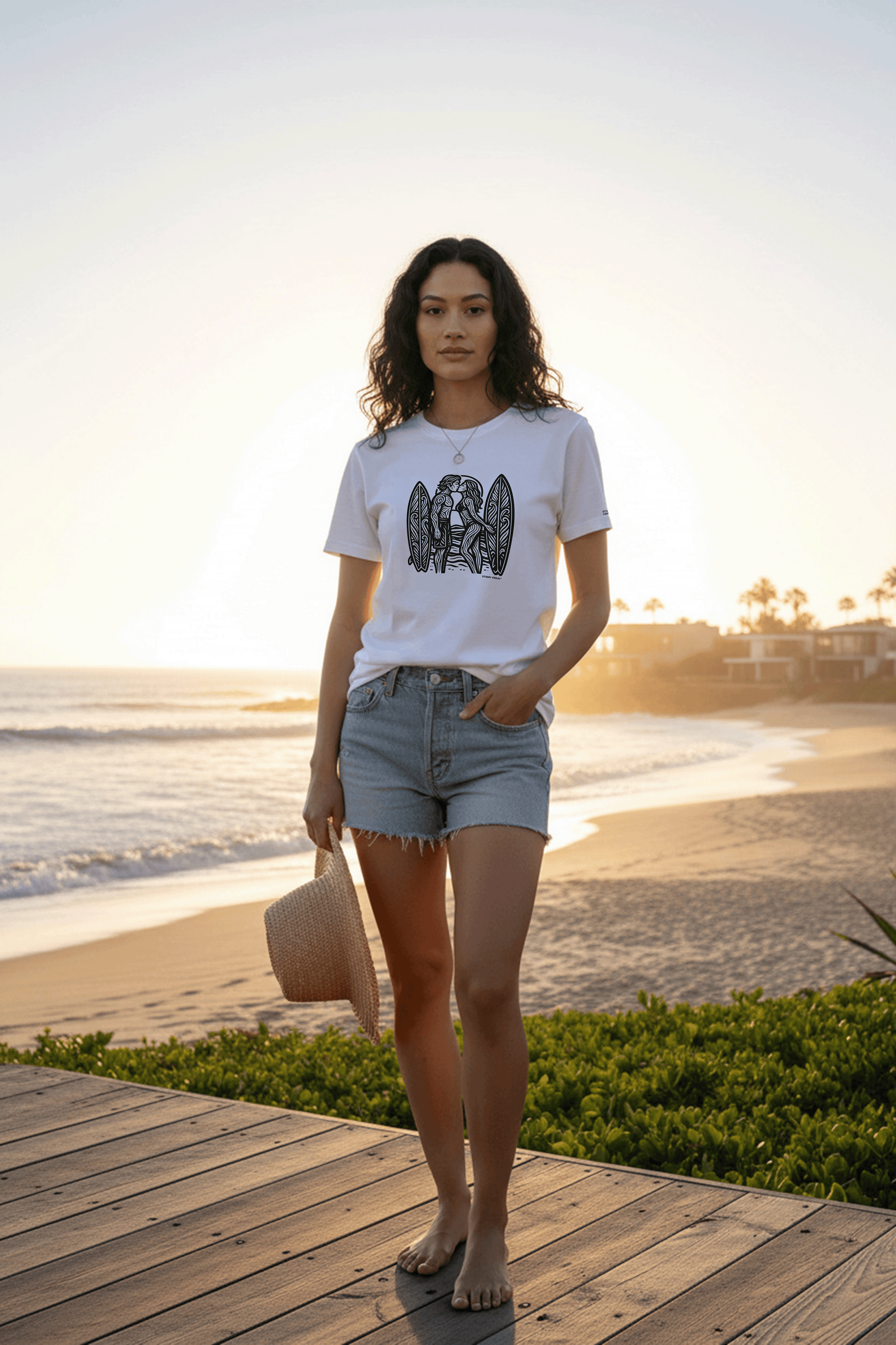 Model standing on a wooden deck by the beach at sunset, wearing a graphic tee by Studio Viekay and denim shorts.