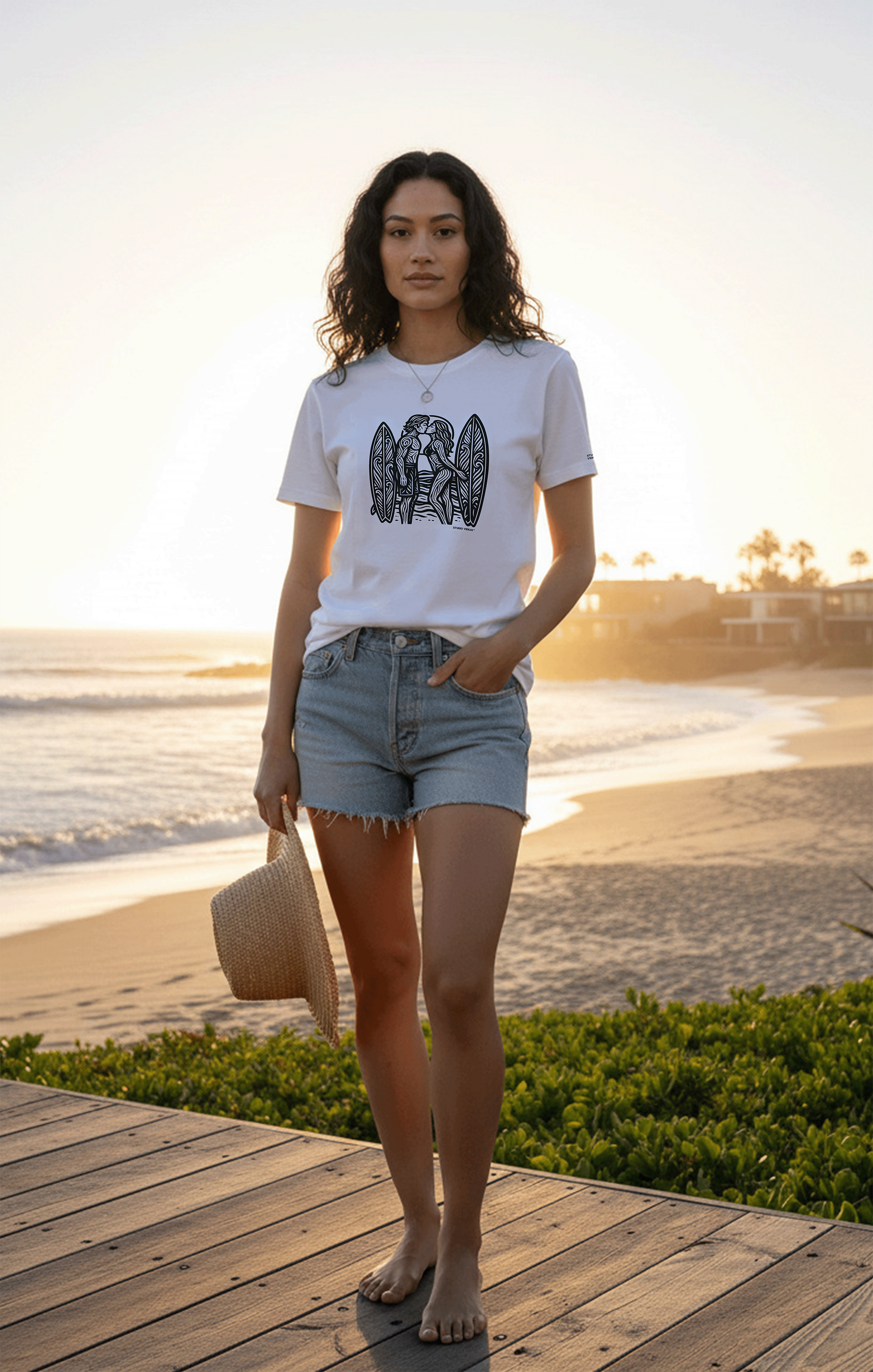 Model standing on a wooden deck by the beach at sunset, wearing a graphic tee by Studio VieKay and denim shorts.